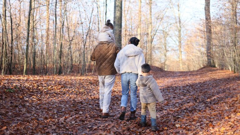A family with two young children walking along a leaf-covered forest path in autumn